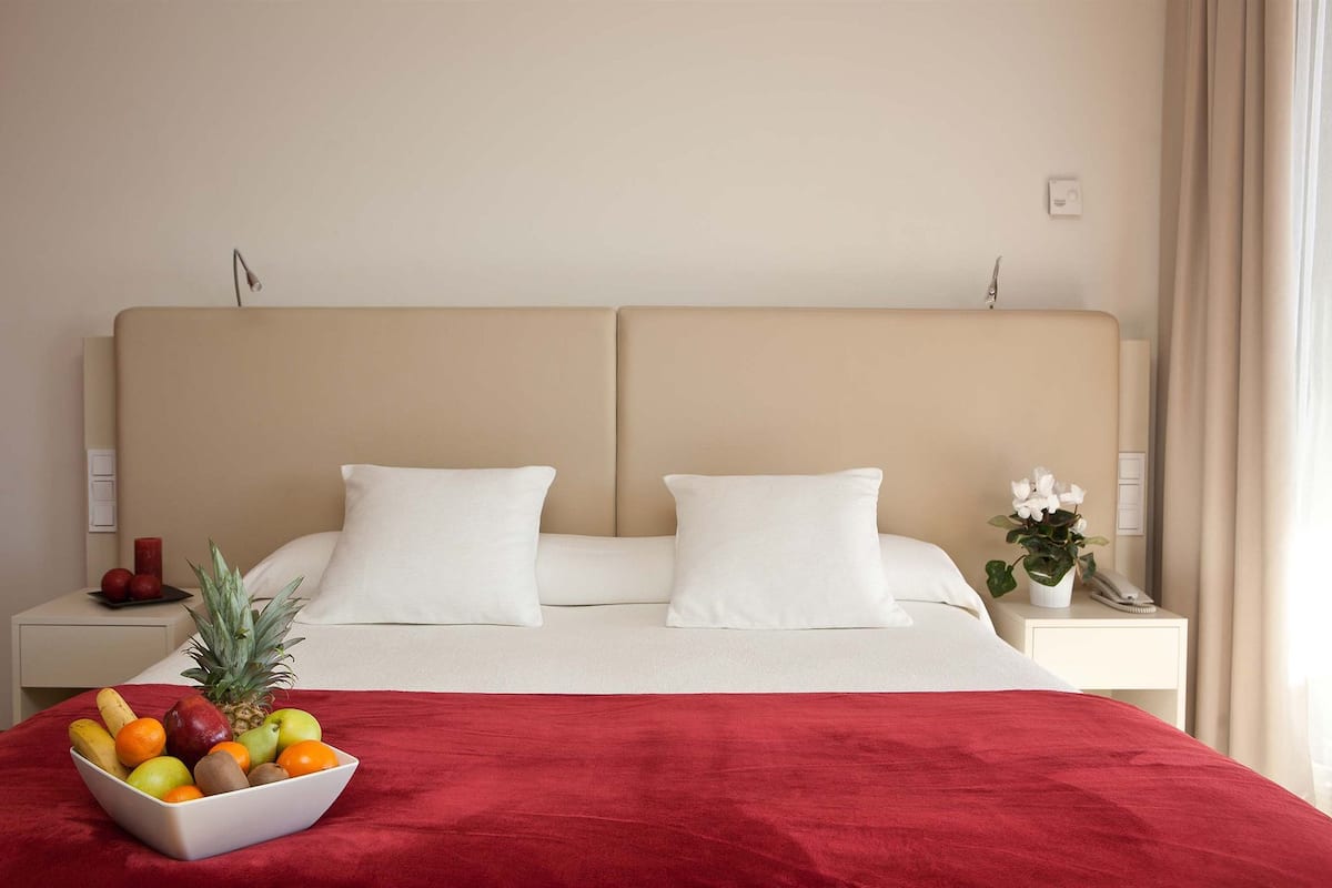 A hotel room with a large bed, two pillows, a nightstand with a phone and a plant, and a fruit bowl on the side table.