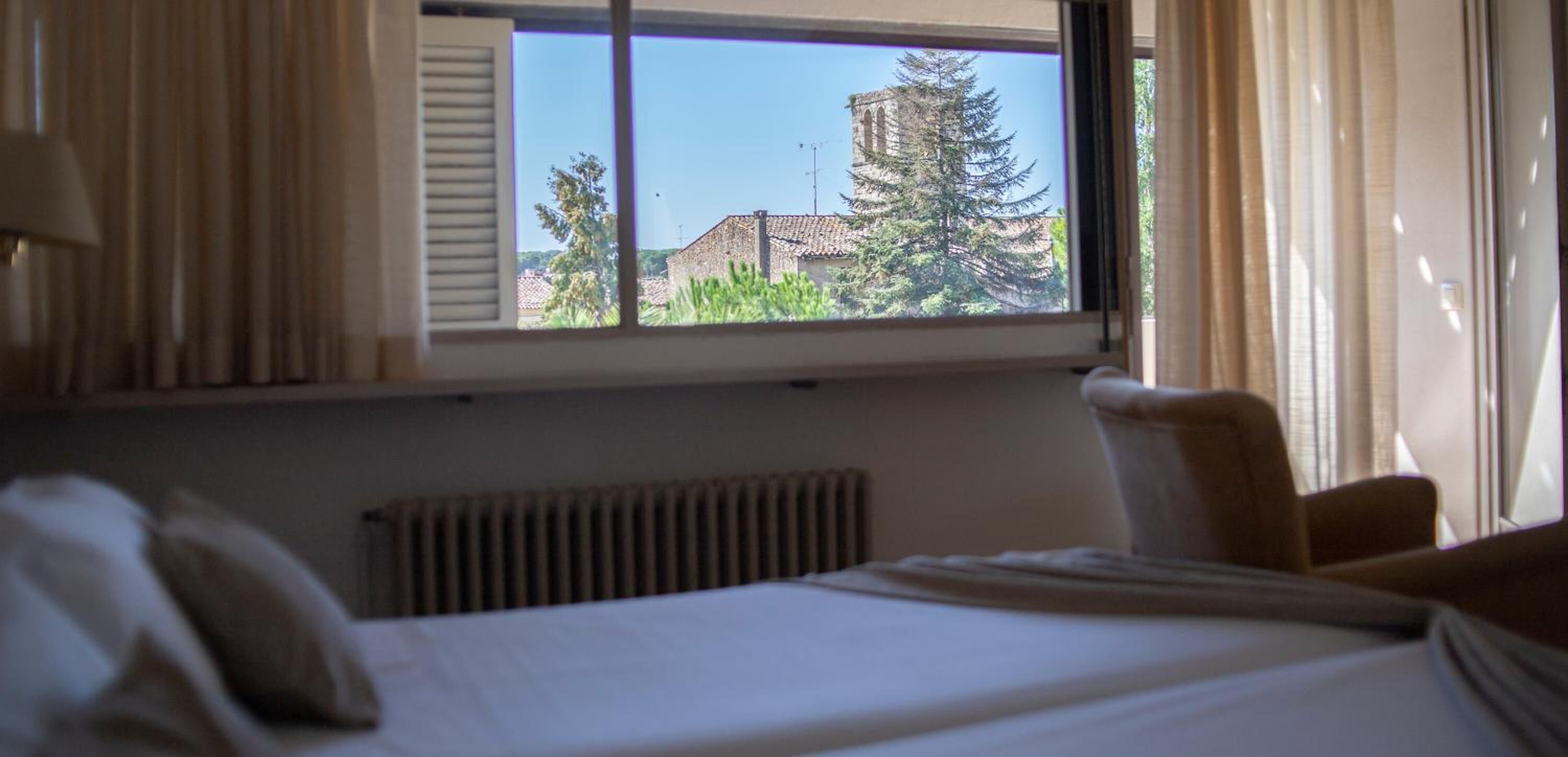 Quarto de hotel com cama, janela com cortinas e vista para um edifício e árvores lá fora.