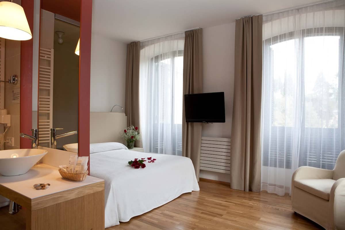 A hotel room with a bed, a TV, a bathroom with a sink and mirror, and large windows with curtains.