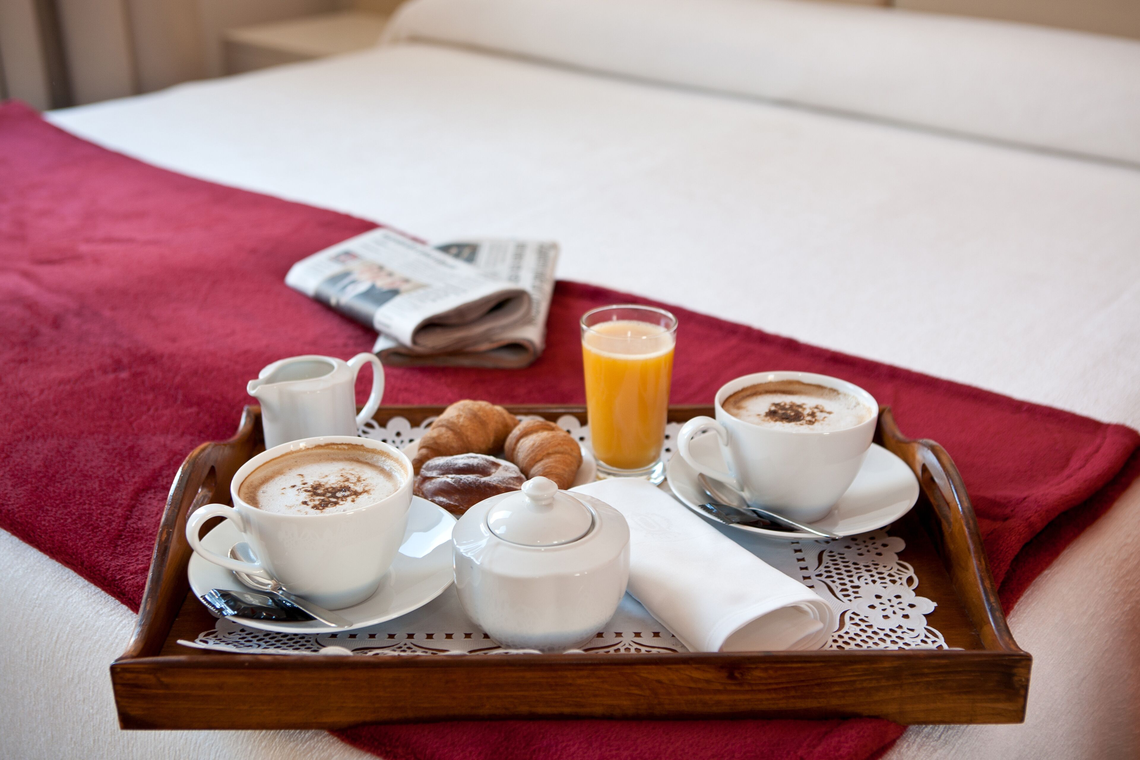 Bandeja de desayuno con cruasanes, café, jarrón de leche y vaso de jugo de naranja sobre una cama con manta roja.