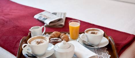 Bandeja de desayuno con cruasanes, café, jarrón de leche y vaso de jugo de naranja sobre una cama con manta roja.