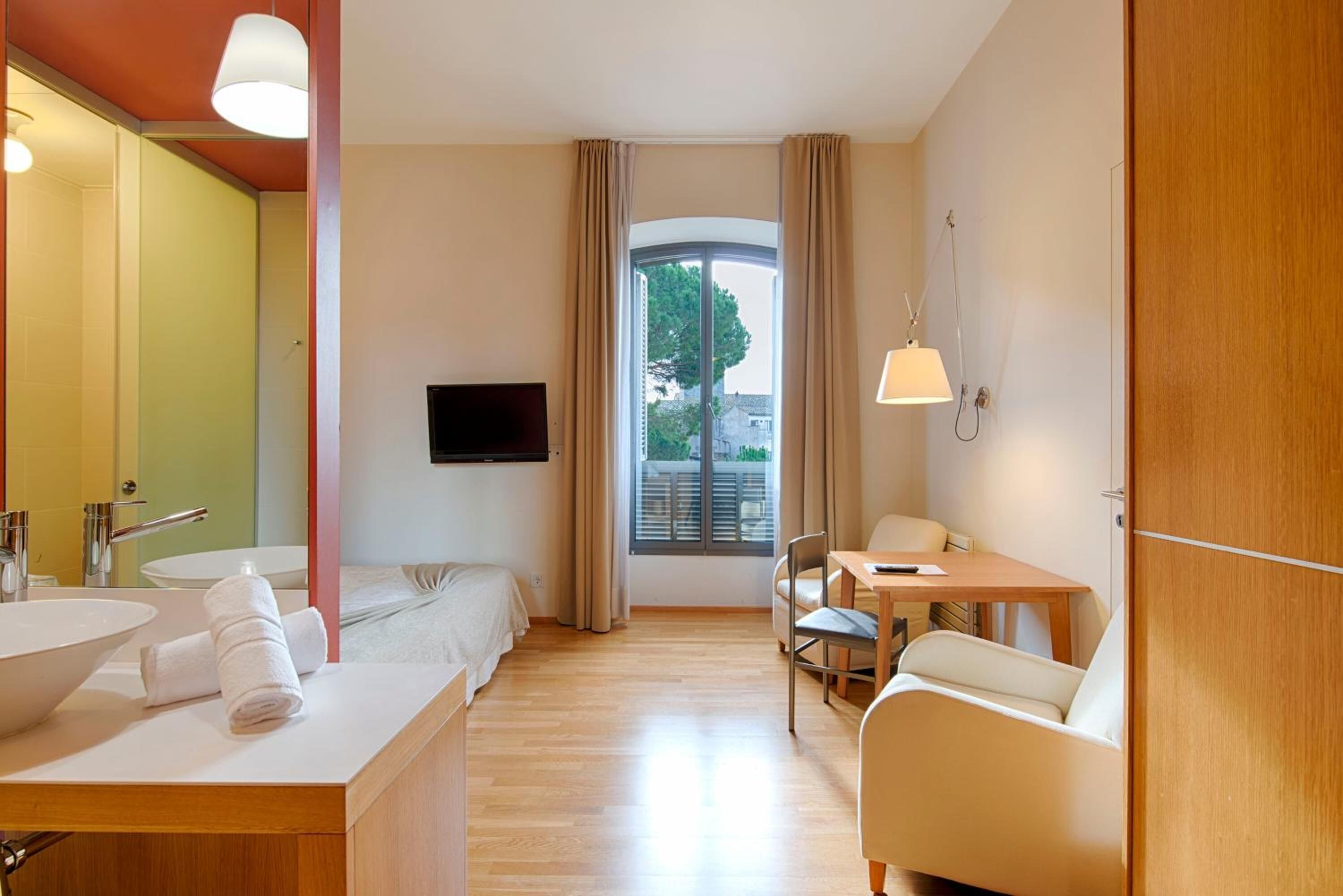 Une chambre d’hôtel dotée d’une grande fenêtre, d’un lit, d’un bureau avec une chaise, d’une télévision et d’une salle de bain avec un lavabo et un miroir.
