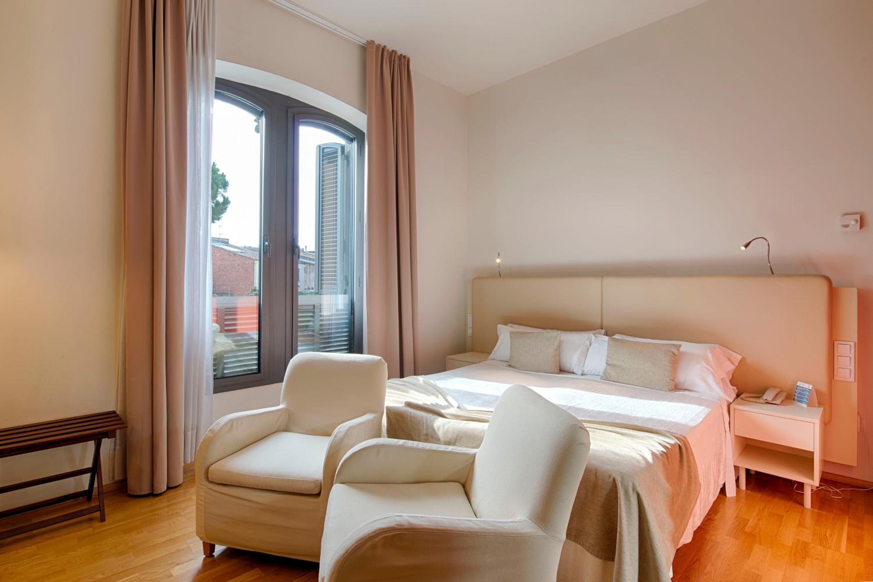 Habitación de hotel con una cama grande, dos sillones, una mesita de noche y una ventana con cortinas.
