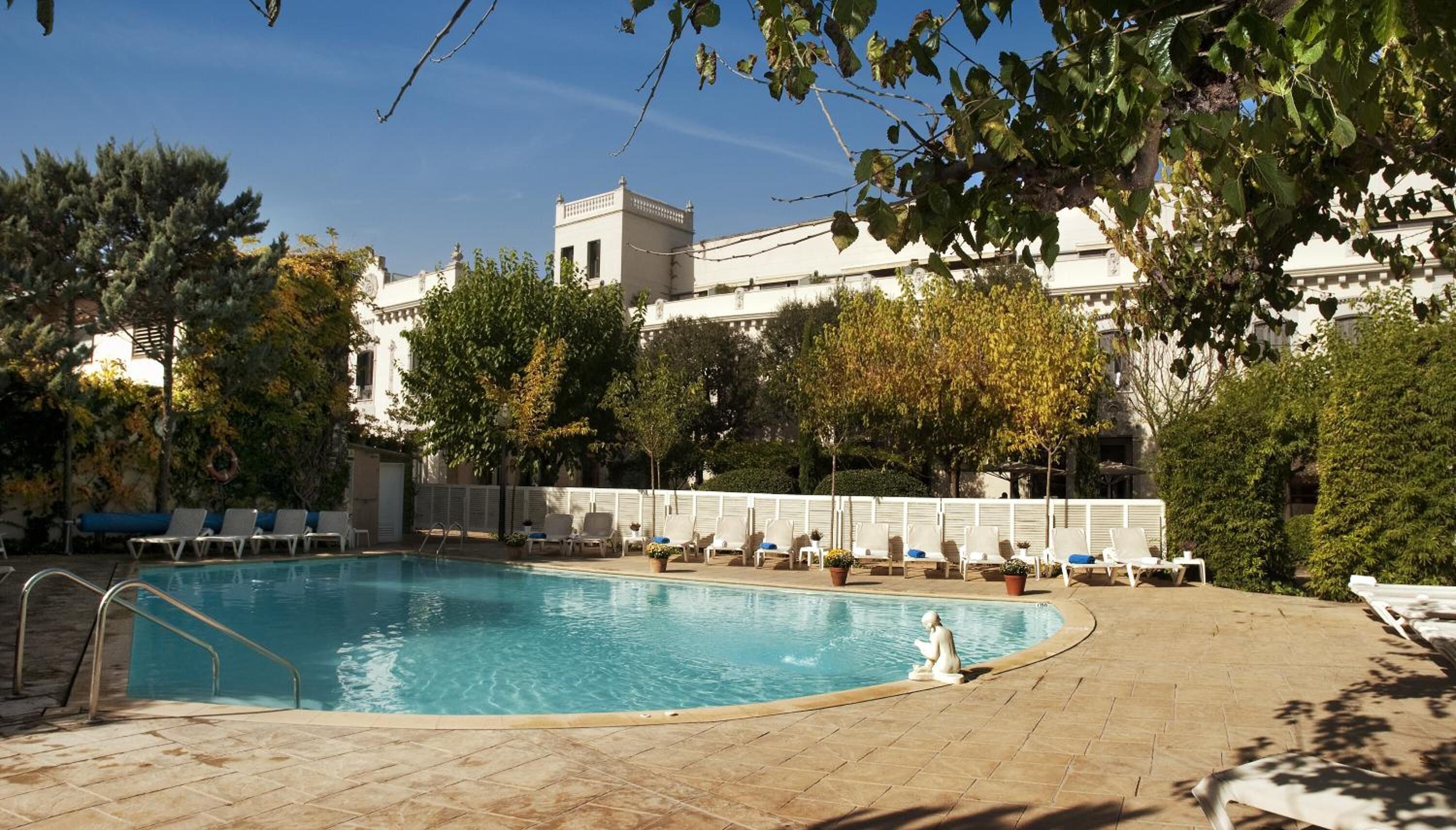 A swimming pool with lounge chairs and a statue, surrounded by trees and buildings.
