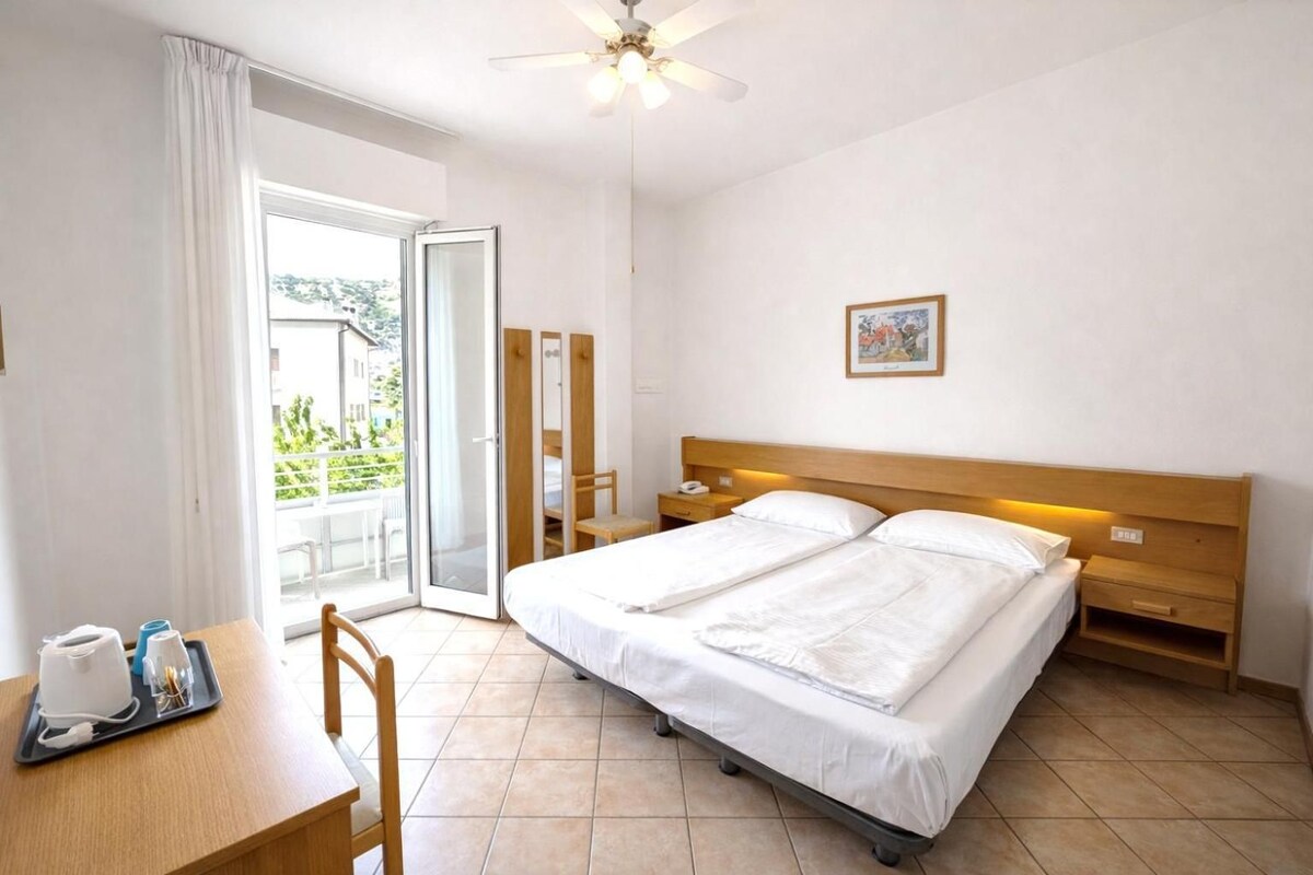 A hotel room with a bed, a desk with a chair, a bedside table, a ceiling fan, and a view through a glass door.