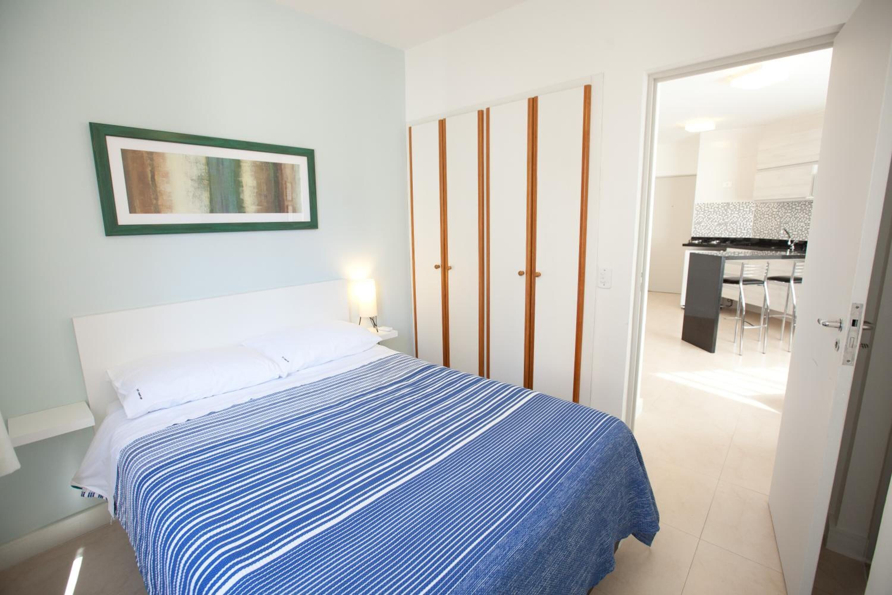 Quarto com cama, guarda-roupa e uma kitchenette ao fundo.