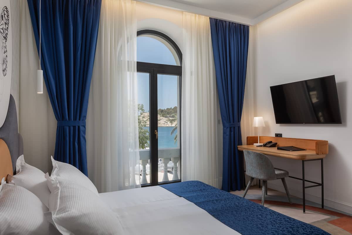 Een hotelkamer met een bed, een bureau, een stoel, een televisie en een balkon met uitzicht op zee.