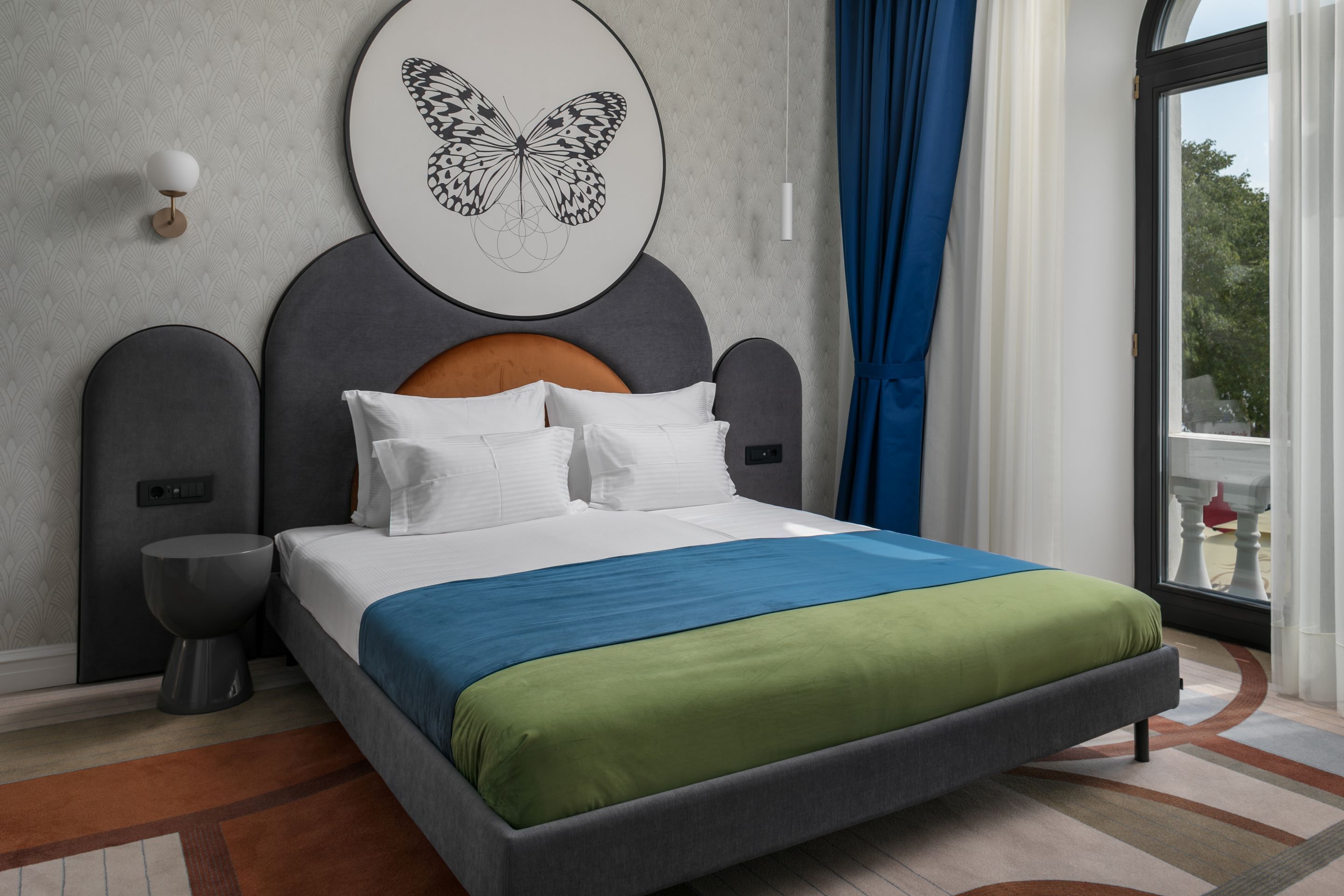 Une chambre d’hôtel moderne avec un grand lit, une tête de lit ornée d’une œuvre d’art représentant un papillon, une table de chevet et une vue sur l’extérieur.