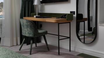 A room with a wooden desk, a green patterned chair, a round mirror, a black telephone, and a lamp.
