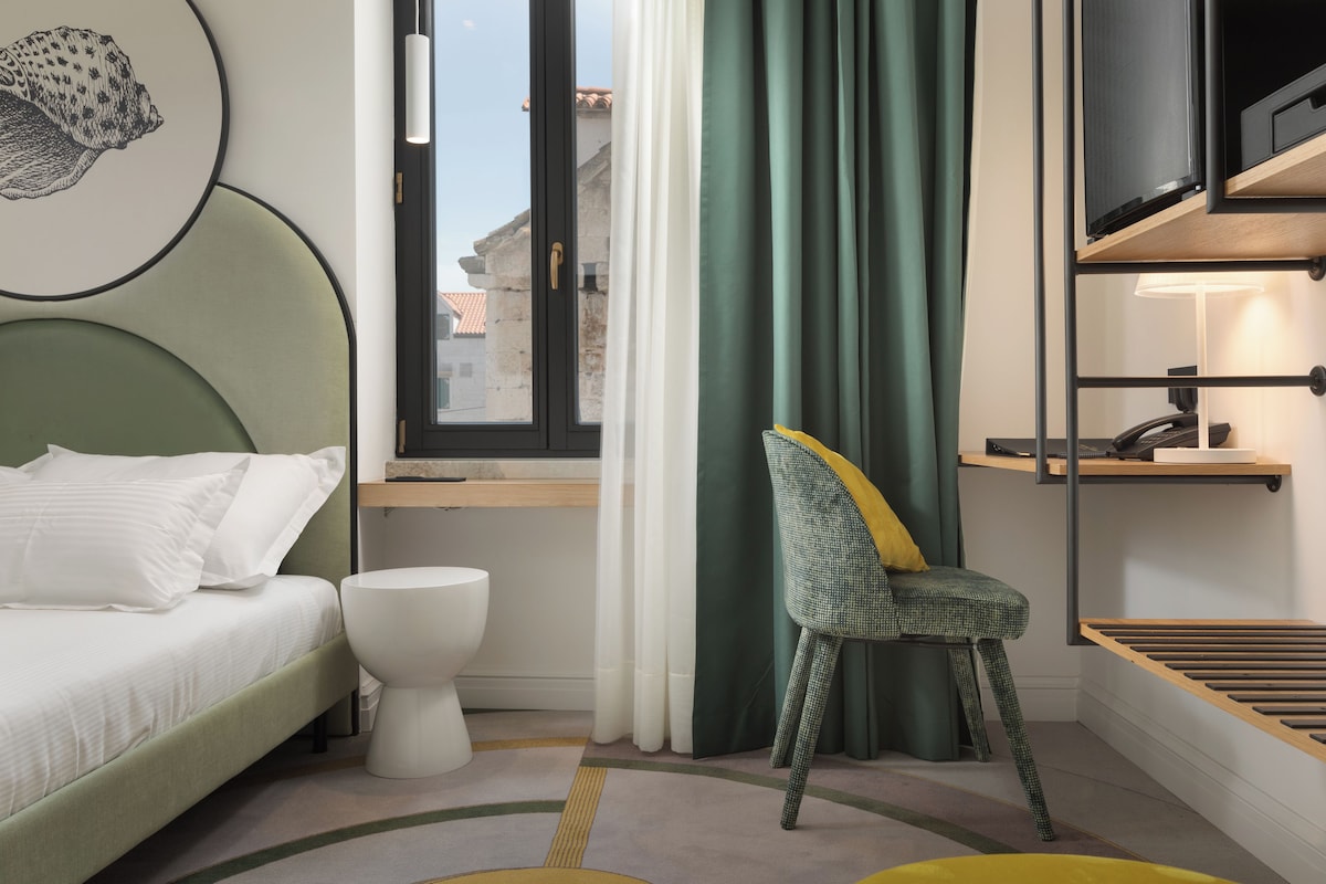 A modern hotel room with a bed, a chair, a toilet, and a window with curtains.