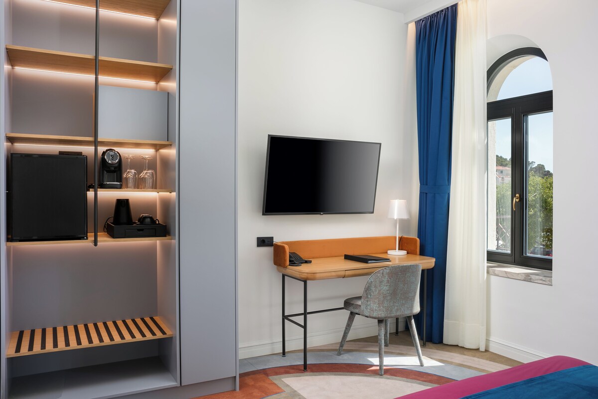 A modern hotel room with a flat-screen TV mounted on the wall, a wooden desk with a chair, a small shelf with a coffee maker, and a window with blue curtains.