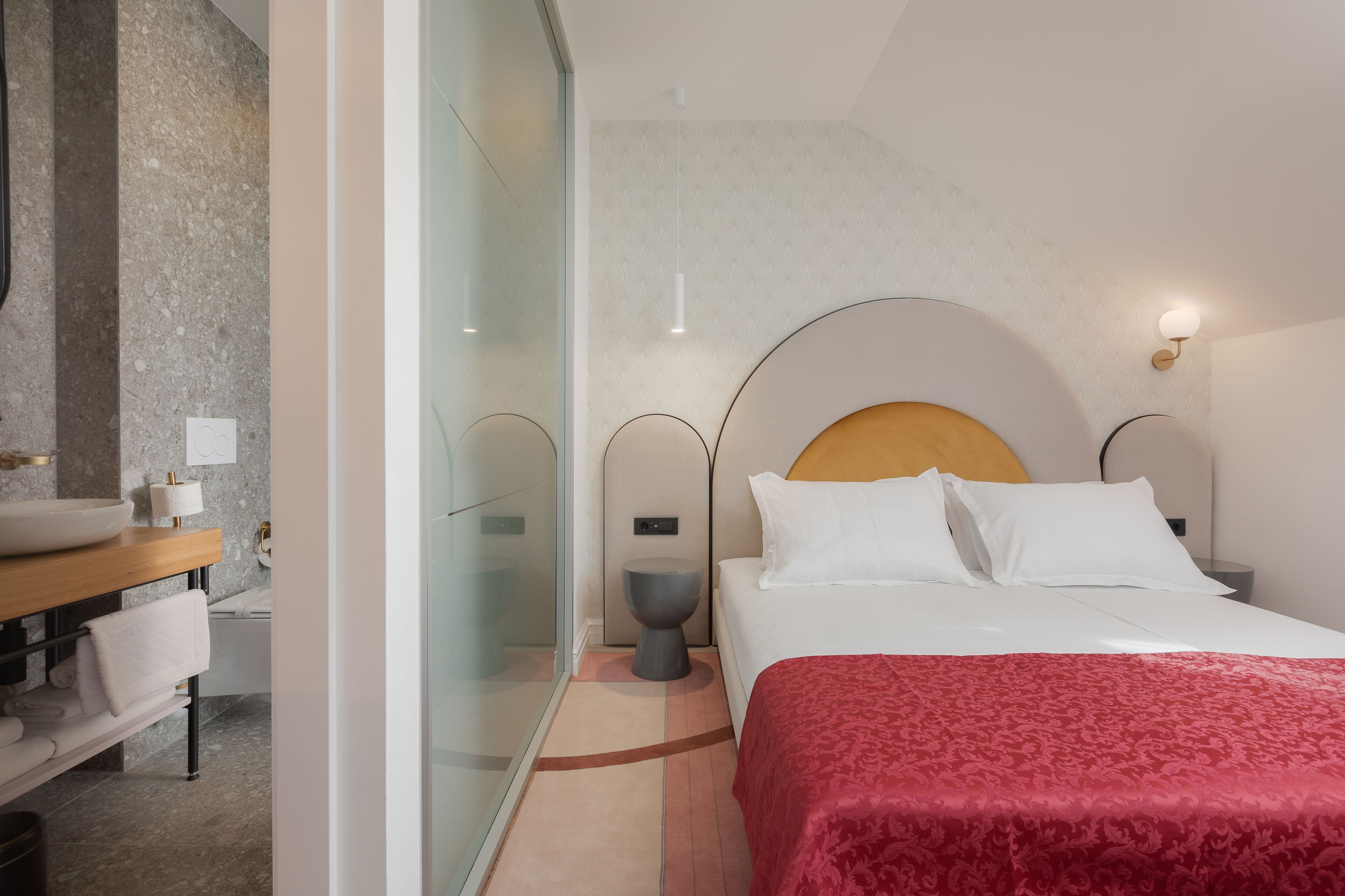 A modern hotel room with a large bed, a wooden headboard, and a glass partition separating the bedroom from the bathroom.