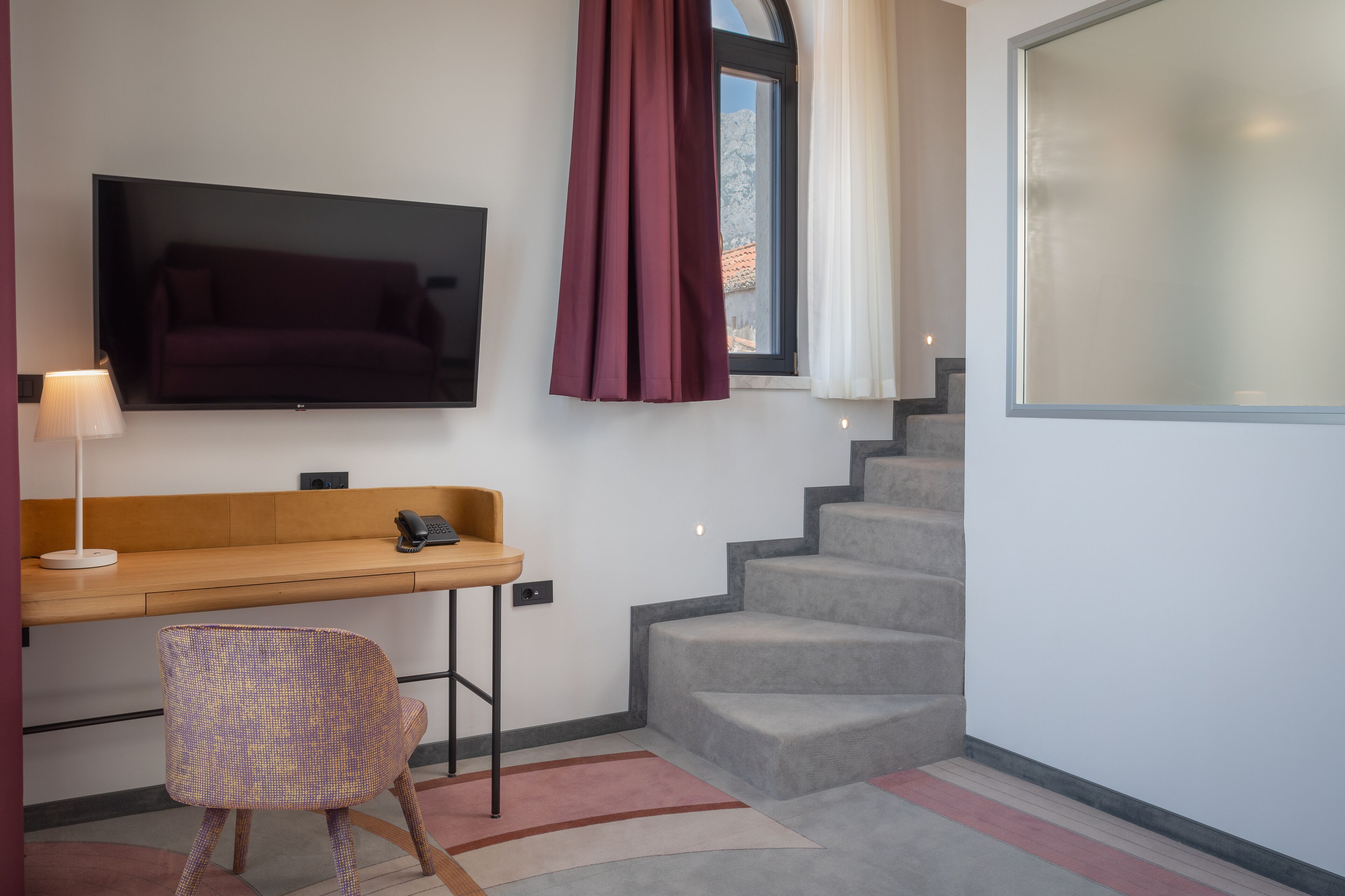 A modern hotel room with a flat-screen TV mounted on the wall, a wooden desk with a chair, a staircase, and a large window with curtains.