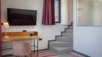 A modern hotel room with a flat-screen TV mounted on the wall, a wooden desk with a chair, a staircase, and a large window with curtains.