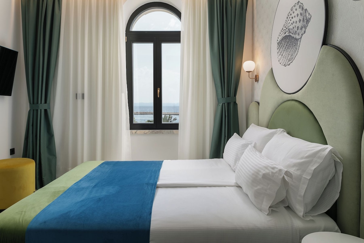 Quarto de hotel com uma cama, vista para o mar através de uma janela e uma obra de arte na parede.