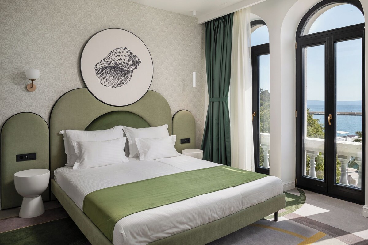 Een hotelkamer met een bed, een groen hoofdeinde, een kunstwerk aan de muur, uitzicht op zee en een balkon met een hekwerk.