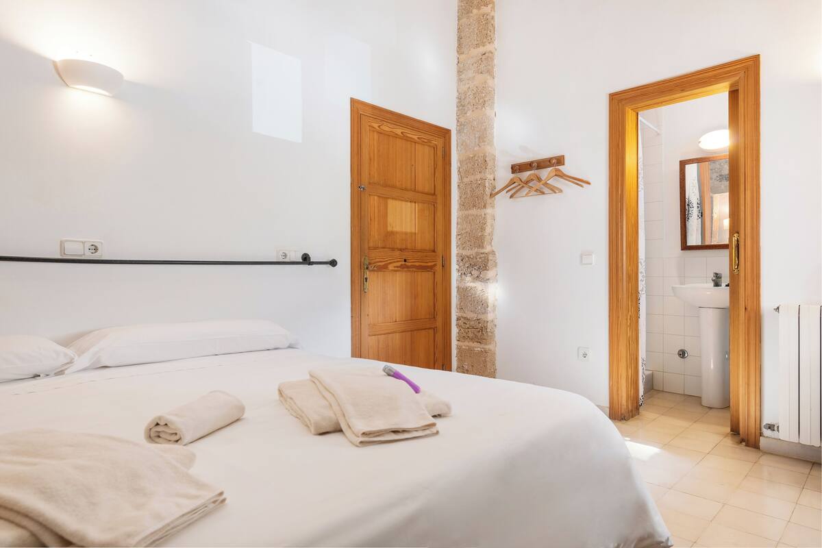 Ein Hotelzimmer mit Bett, Handtüchern, einer Holztür, einer Steinsäule, einem spiegelnden Rahmen aus Holz und einem Badezimmer mit Waschbecken und Handtuchhalter.