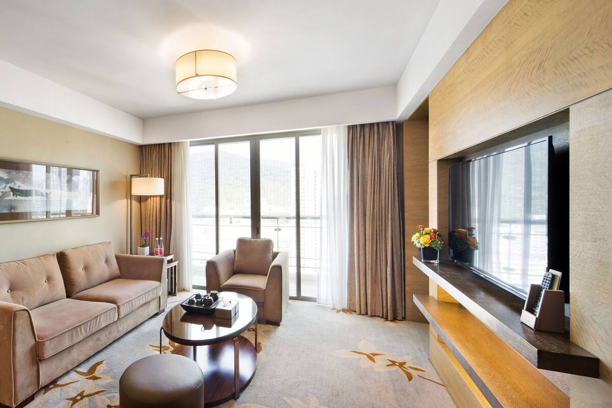 A modern hotel room with a sofa, armchair, and coffee table, large windows with curtains, and a city view.