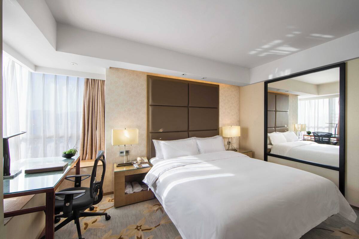 A hotel room with a large bed, a desk with a chair, a small table with a lamp, and a large mirror.
