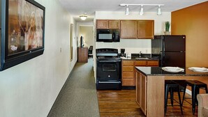 A modern kitchen with wooden cabinets, a black refrigerator, a microwave, and a stove. There is a bar area with stools and a television mounted on the wall. - Affordable Suites Augusta (Augusta)