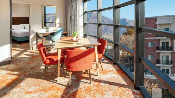 A hotel room with a large window, a bed, a desk, and a dining area with chairs. - Hilton Garden Inn Ogden UT (Ogden)