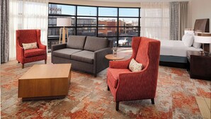 A hotel room with a large window, a grey sofa, a red armchair, a wooden coffee table, and a bed with white bedding. - Hilton Garden Inn Ogden UT (Ogden)