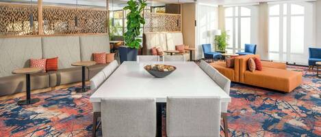 A modern hotel lobby with a long dining table, comfortable seating, and large windows.