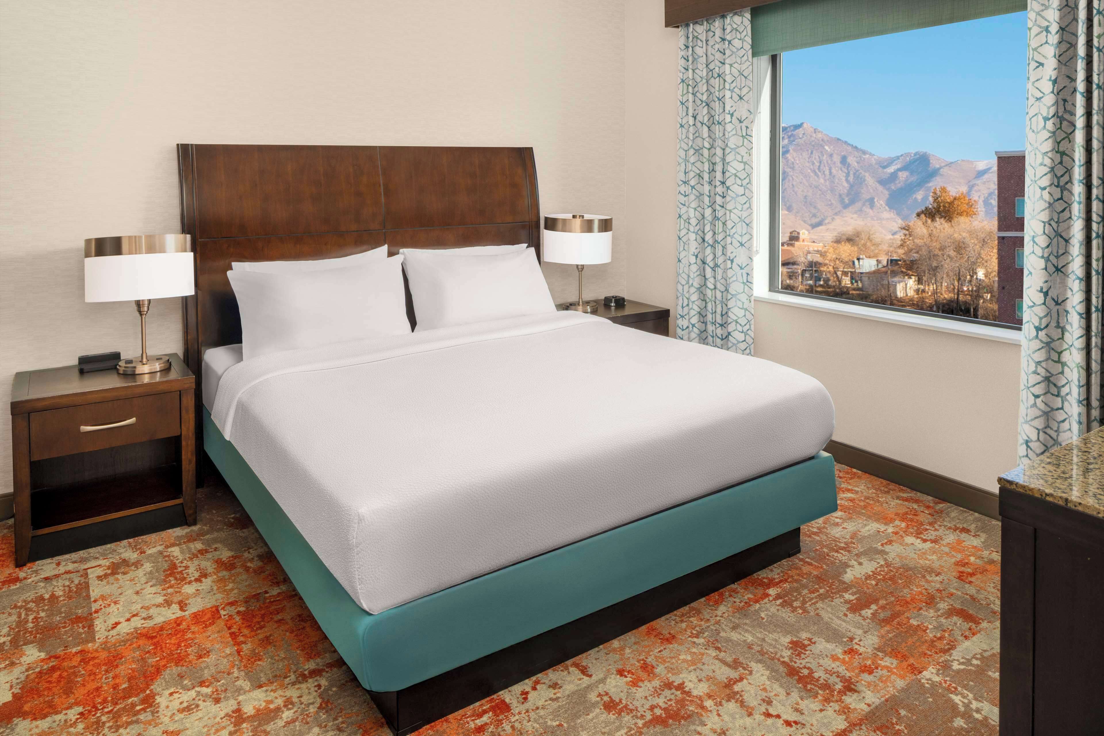 Habitación de hotel con una cama grande, dos mesitas de noche con lámparas, vistas a montañas a través de una ventana y una alfombra estampada.