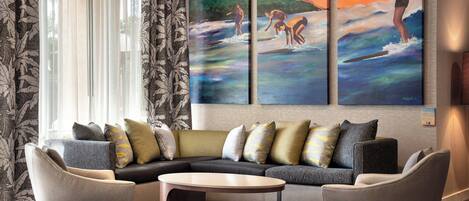 A modern living room with a sectional sofa, two armchairs, and a coffee table. There are three large paintings on the wall depicting surfing scenes.