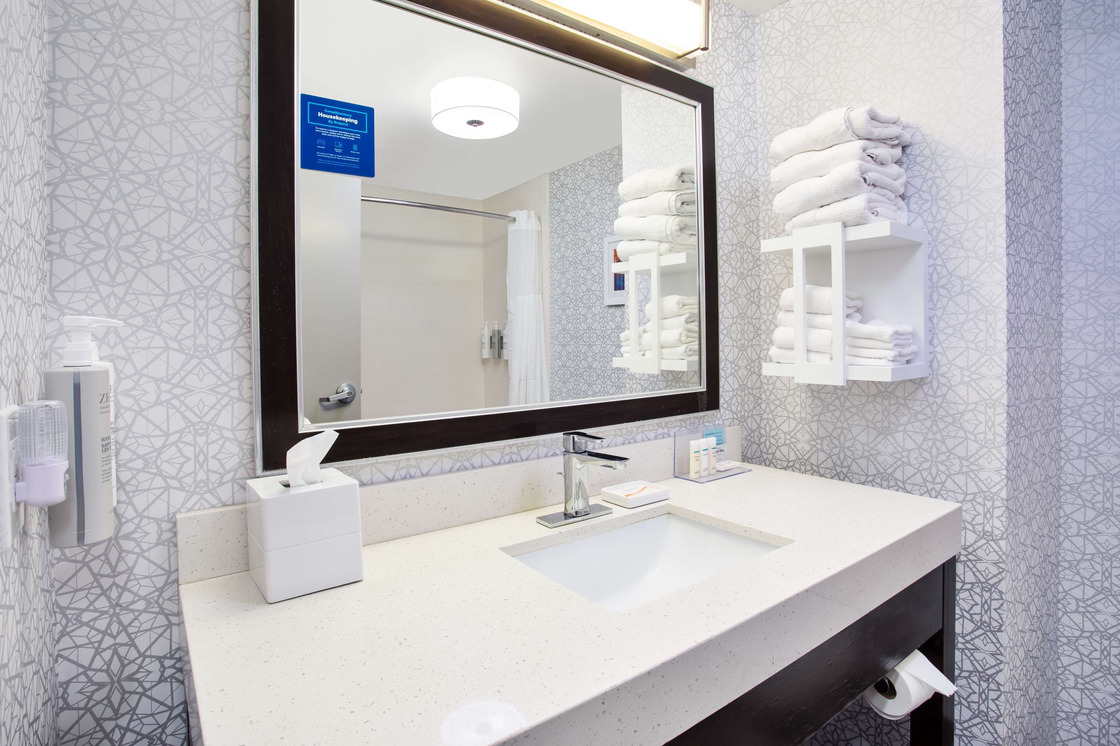 Un baño moderno con un espejo grande, un lavabo, un perchero para toallas y un dispensador de gel antibacterial.
