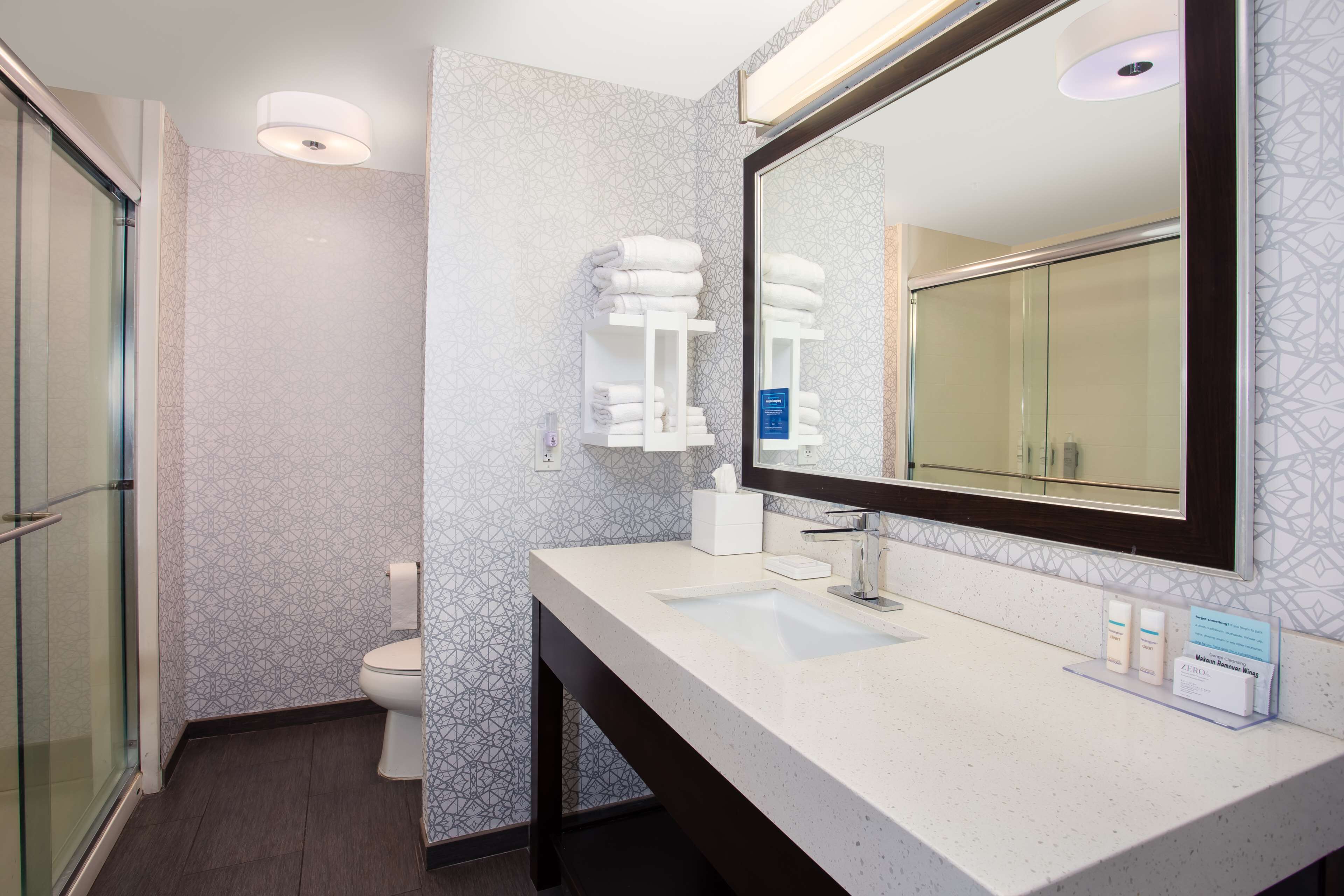 Een moderne badkamer met een douchecabine van glas, een witte wastafel met een grote spiegel en netjes opgevouwen handdoeken.