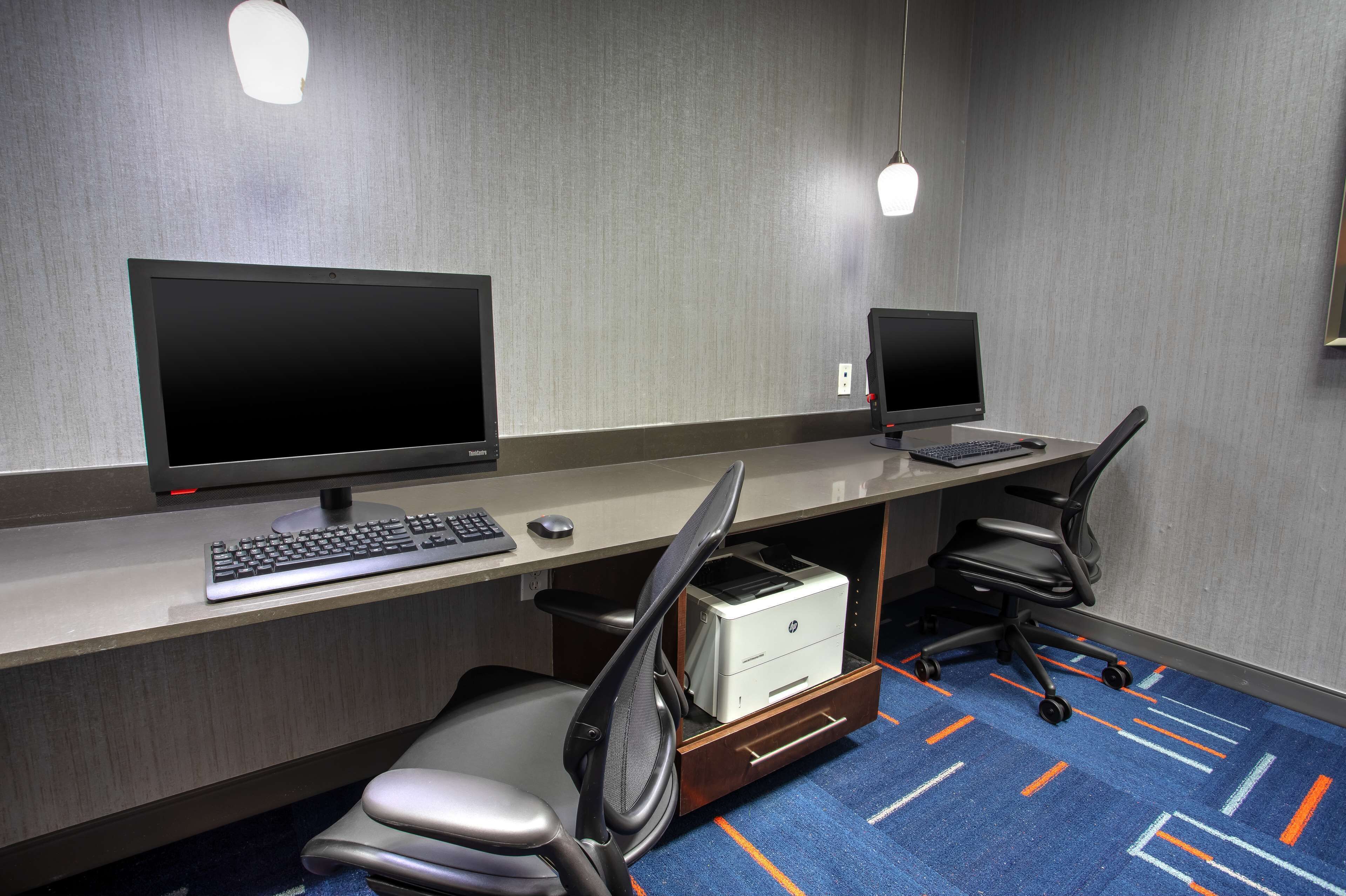 Et hotelkontor med to skriveborde, hver med computerskærm, tastatur og mus. To kontorstole, hvoraf den ene har en skrivebordsorganisator under bordet. Værelset har et tæppe med et geometrisk mønster og en lampe på væggen.