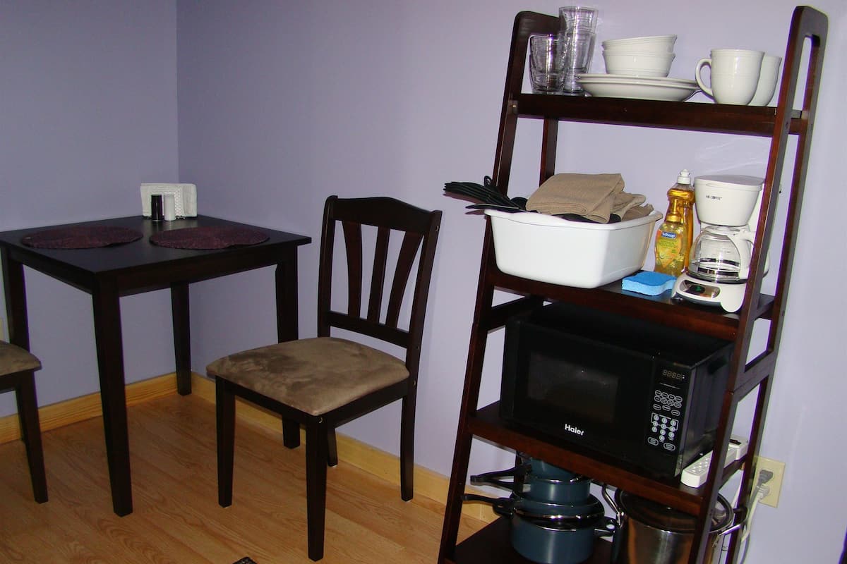A compact kitchen with a wooden table, a chair, a microwave, a coffee maker, and a shelf with dishes and a bottle.