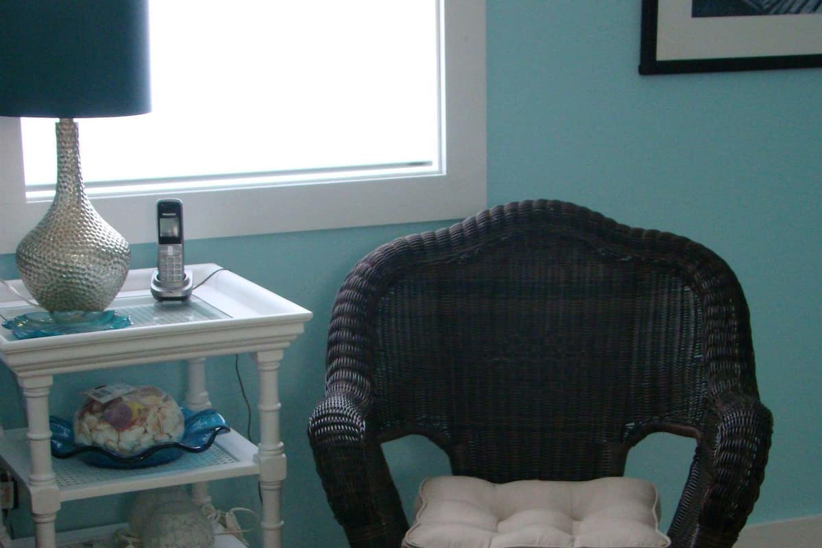 A room with a wicker chair, a green ottoman, a white shelf with decorative items, and a lamp.