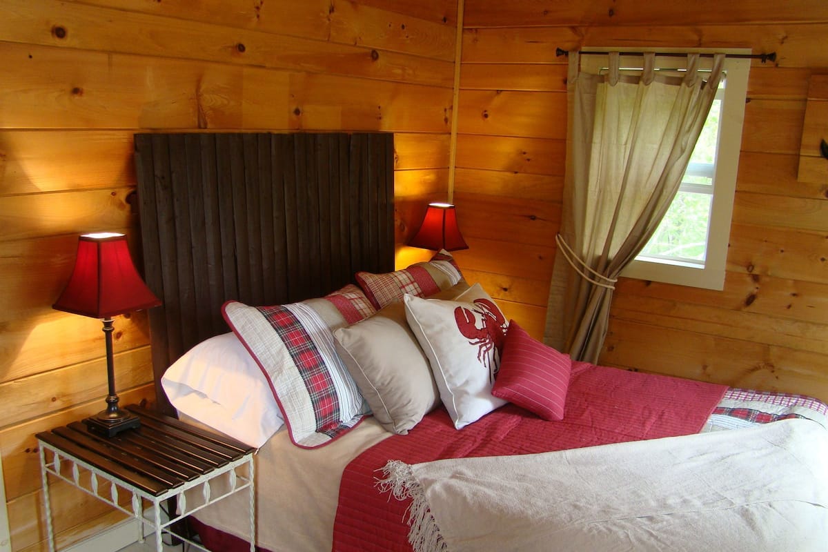 A wooden cabin bedroom with a bed, two bedside lamps, a window with curtains, and a wooden headboard.