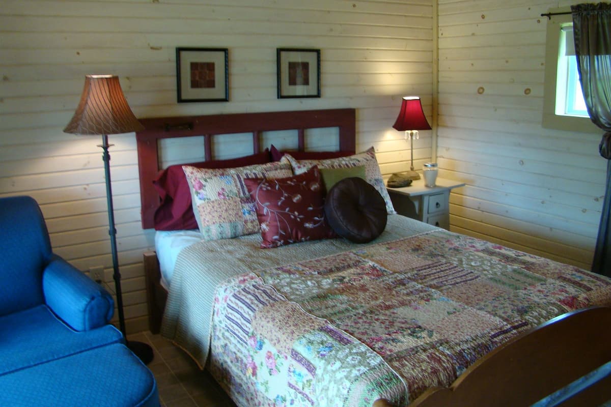 A bedroom with a bed, two bedside lamps, a nightstand, a chair, and wooden walls.