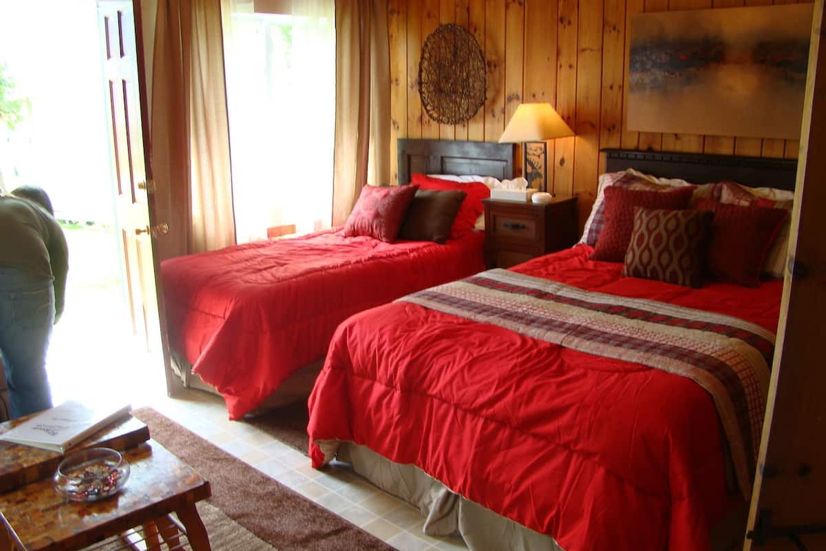 A room with two beds, a wooden headboard, a nightstand, a lamp, and a window with curtains.