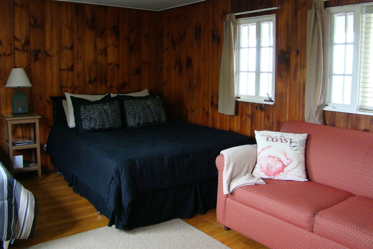 A room with wooden walls, a bed with dark bedding, a red sofa, and a nightstand with a lamp.