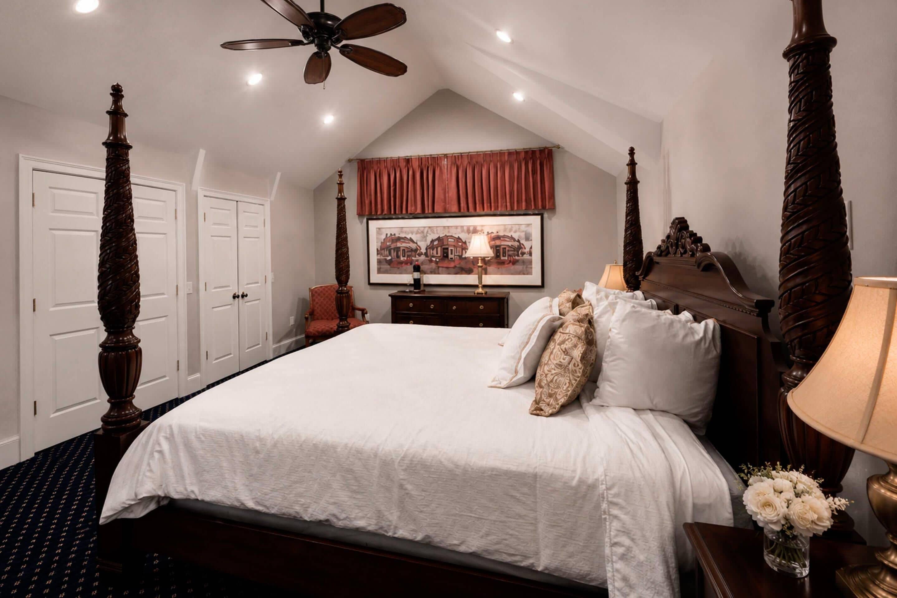 Ein Schlafzimmer mit Himmelbett, Deckenventilator, einem gerahmten Bild an der Wand und einem Nachttisch mit Lampe und Blumen.