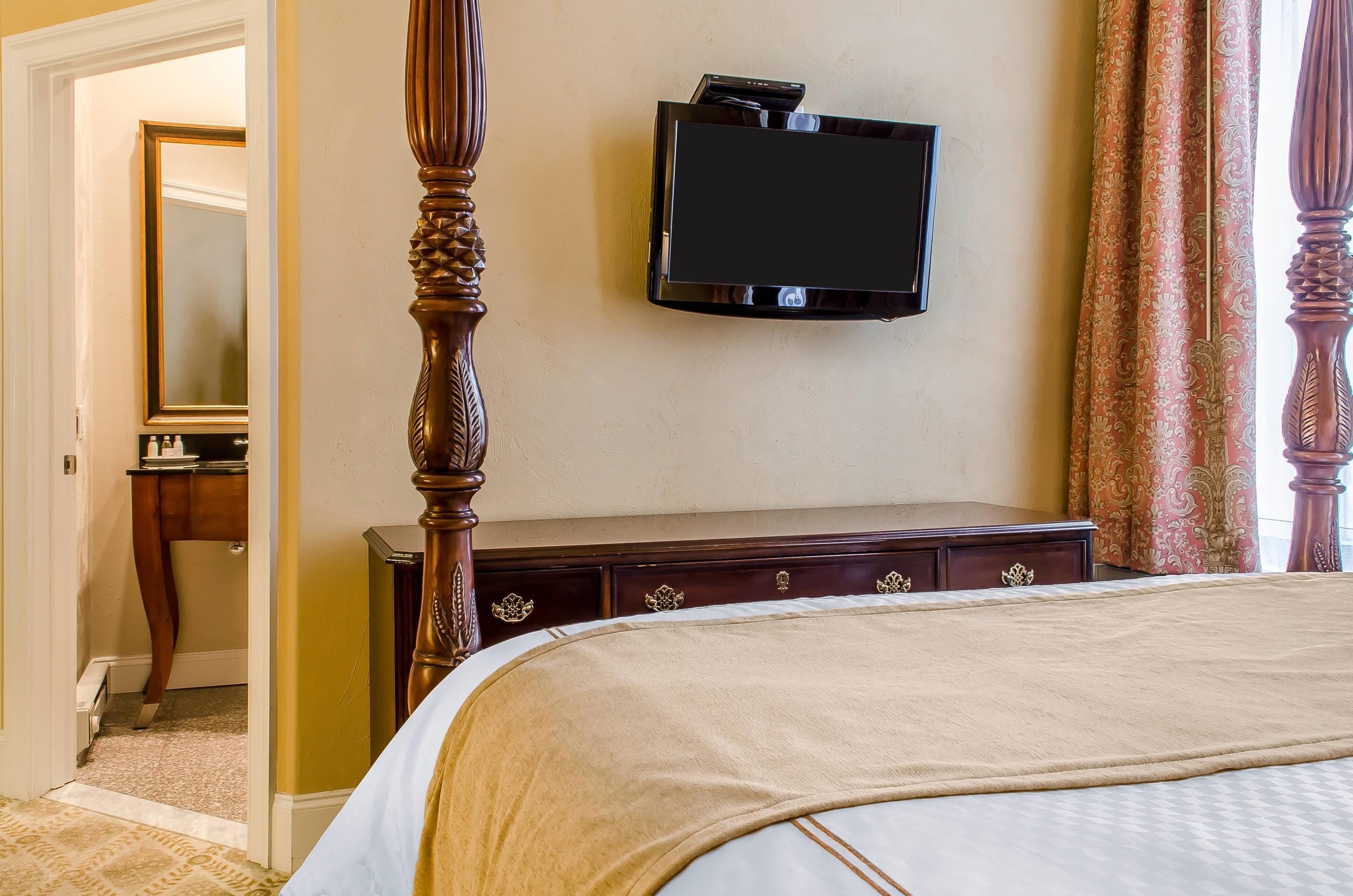 Habitación de hotel con cama con dosel, televisor de pantalla plana en la pared, cómoda de madera con cajones y espejo en una esquina.