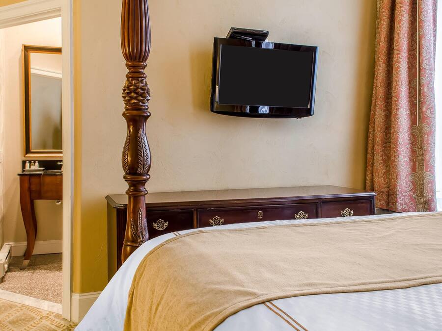 Ein Hotelzimmer mit Himmelbett, Flachbildfernseher an der Wand, einem hölzernen Schminktisch mit Schubladen und einem Spiegel in der Ecke.