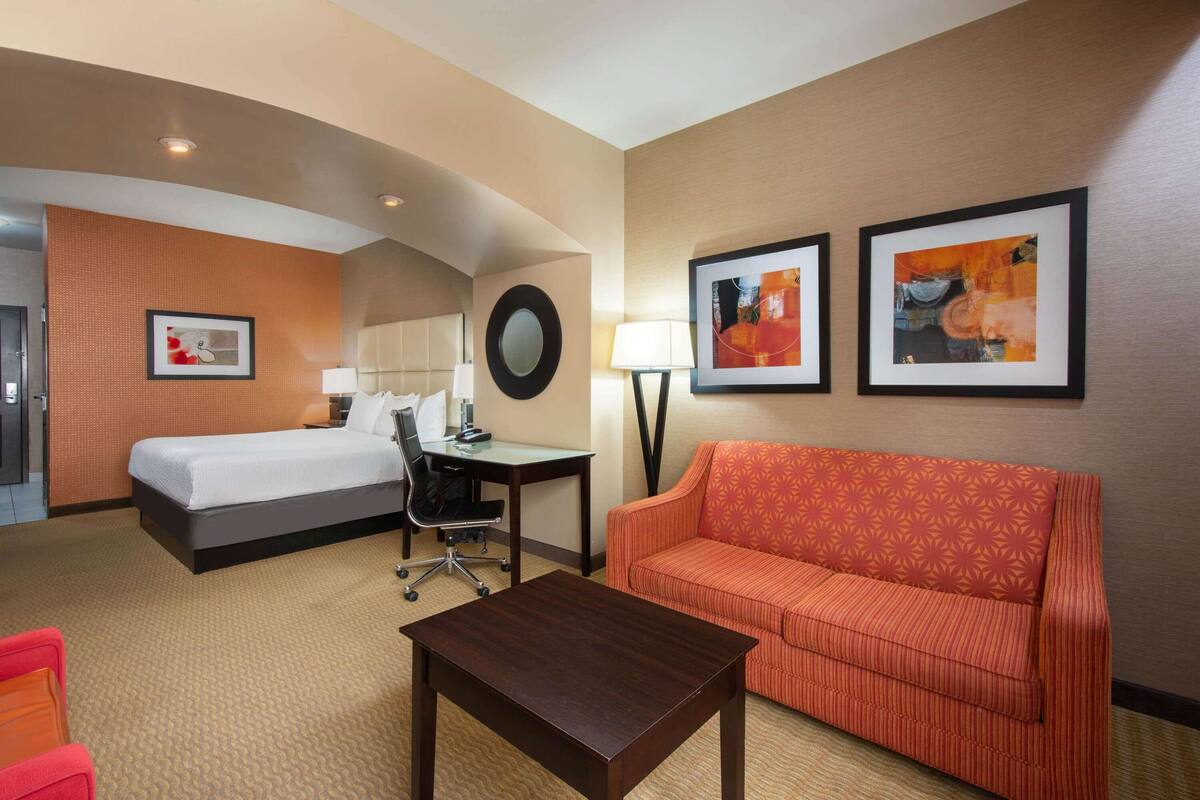 A hotel room with a bed, a desk, a chair, a sofa, and two framed artworks on the wall.