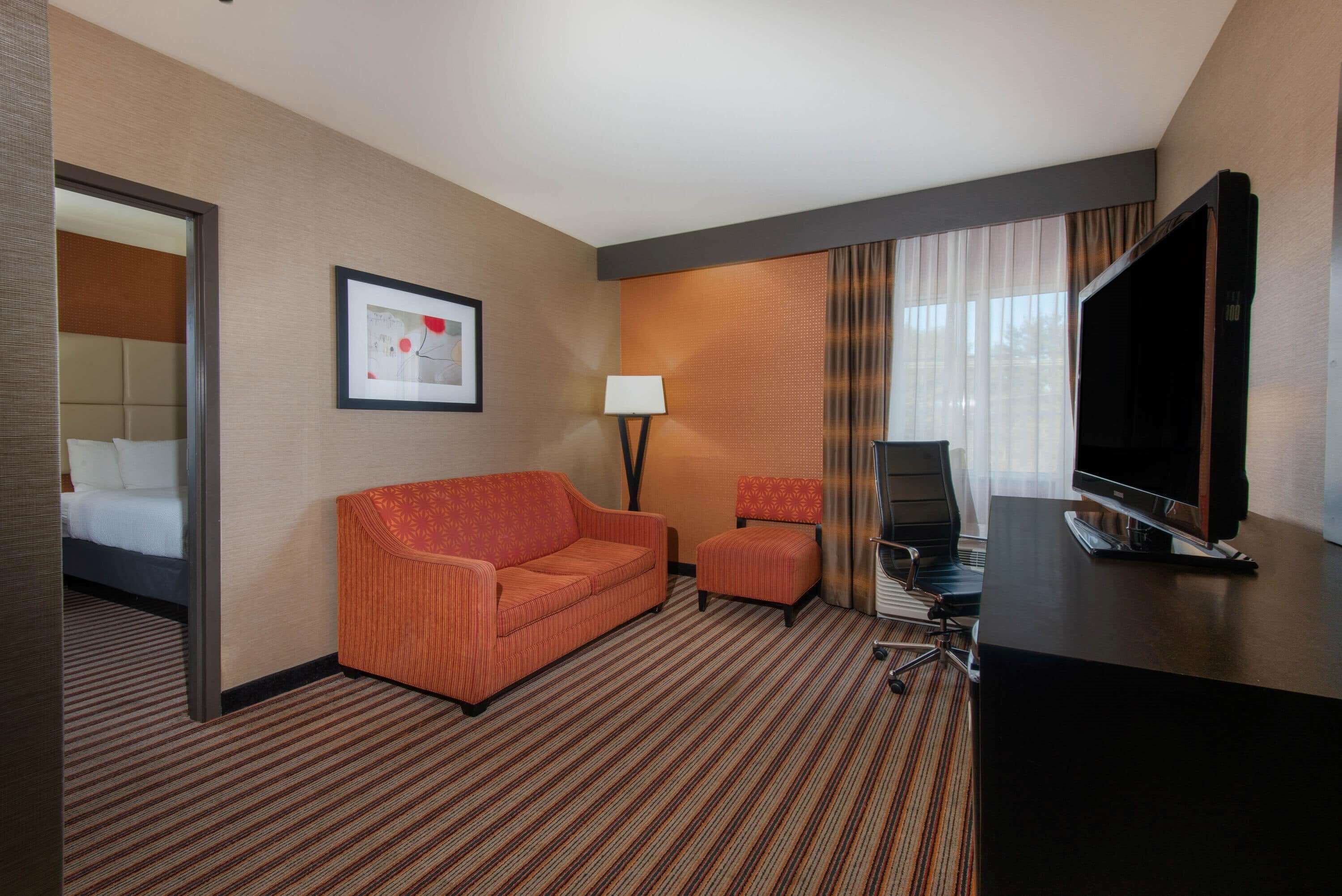 A hotel room with a bed, a sofa, a chair, a desk with a TV, and a lamp.