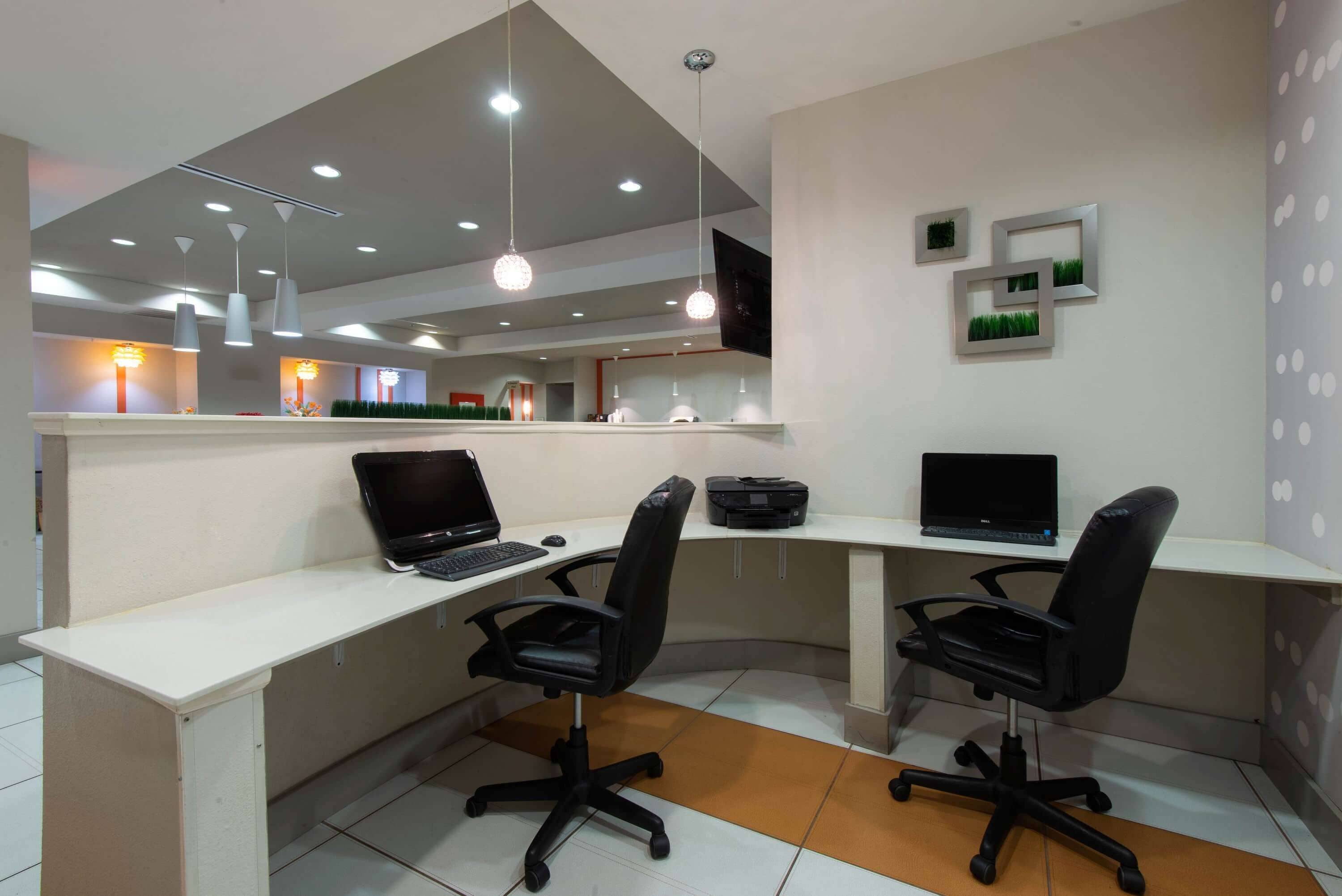 A modern office space with two desks, each with a laptop and a black office chair. There are overhead lights and a flat-screen TV mounted on the wall.