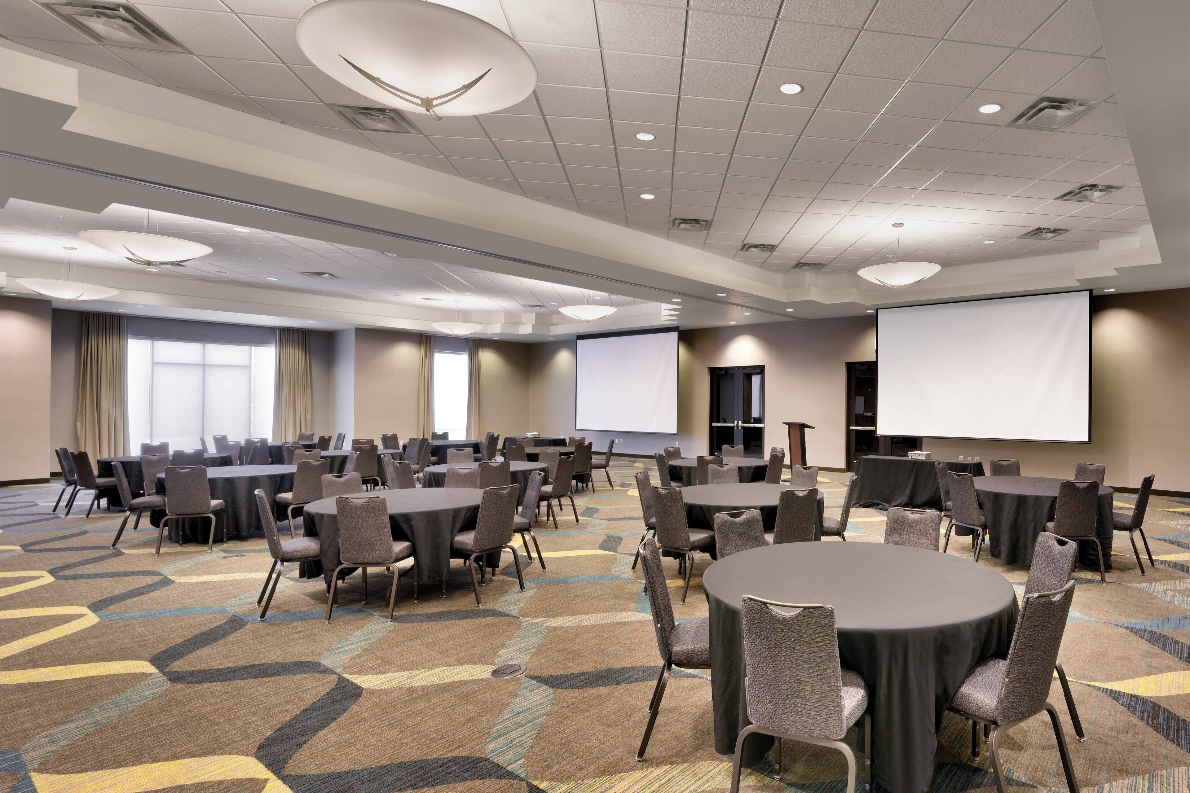 Une salle de conférence équipée de plusieurs tables et chaises, de grandes fenêtres et d’un écran de projection.