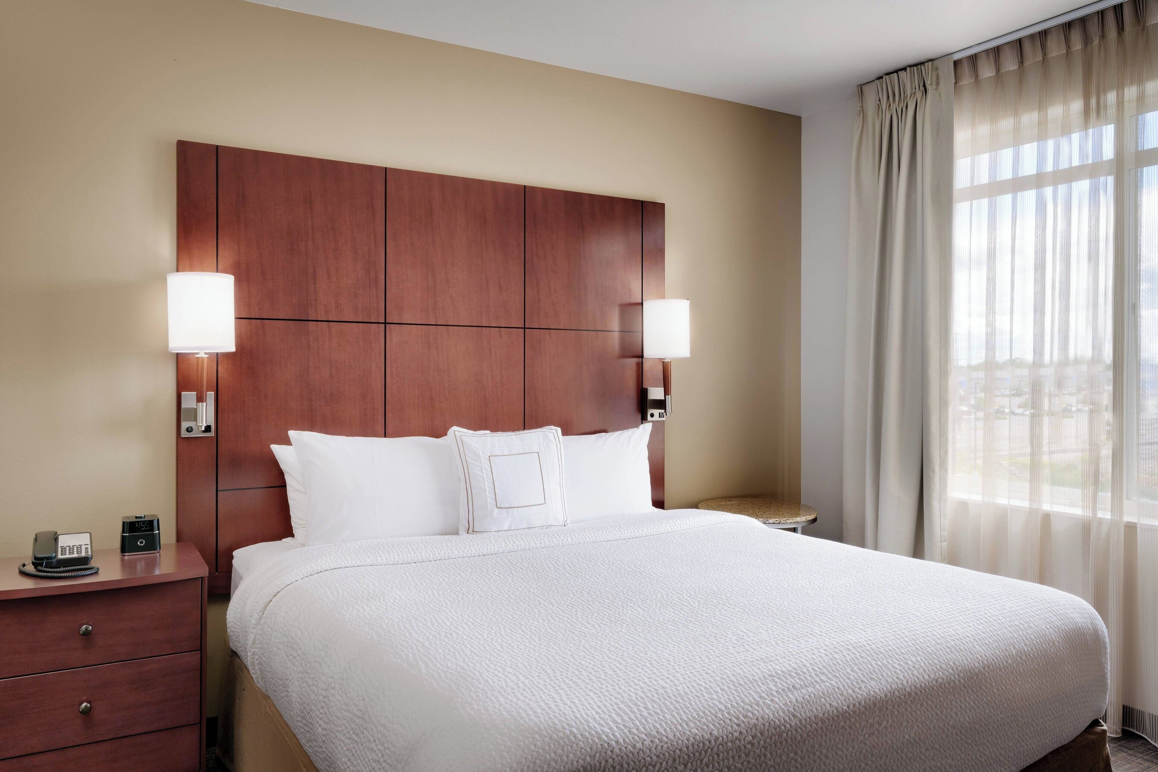 Habitación de hotel con una cama grande, dos mesitas de noche, cabecera de madera y una ventana con cortinas traslúcidas.