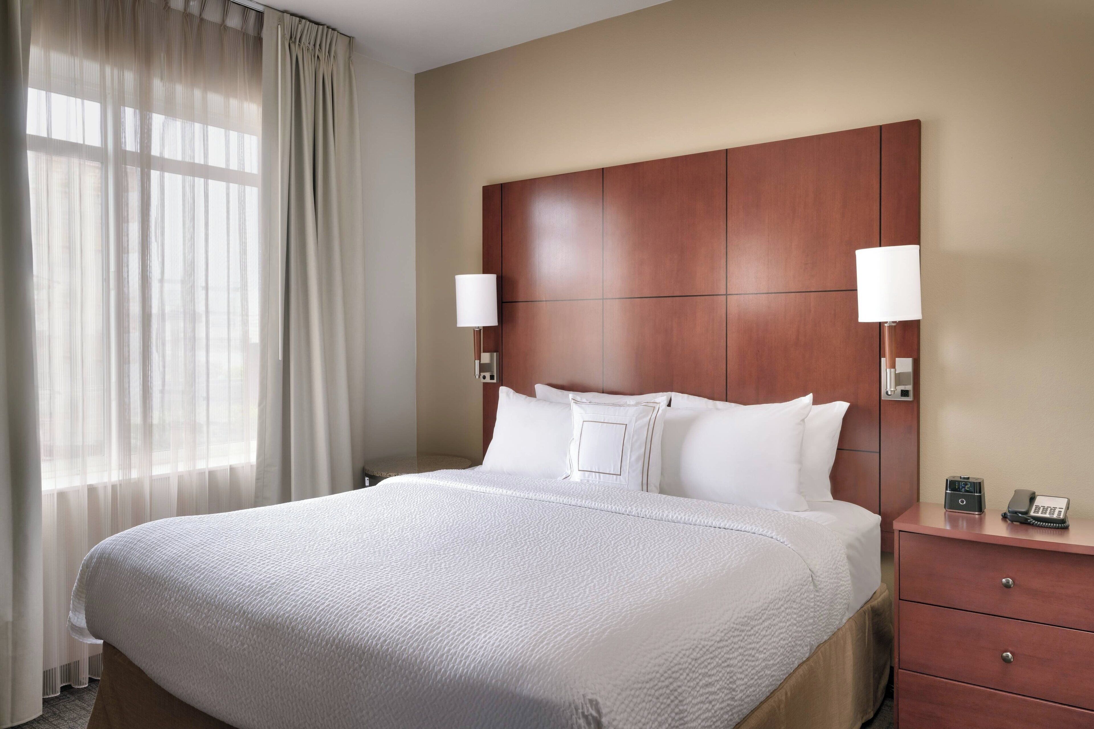 Quarto de hotel com uma cama grande, duas luminárias de cabeceira, uma cabeceira de madeira e uma mesa de cabeceira com telefone.