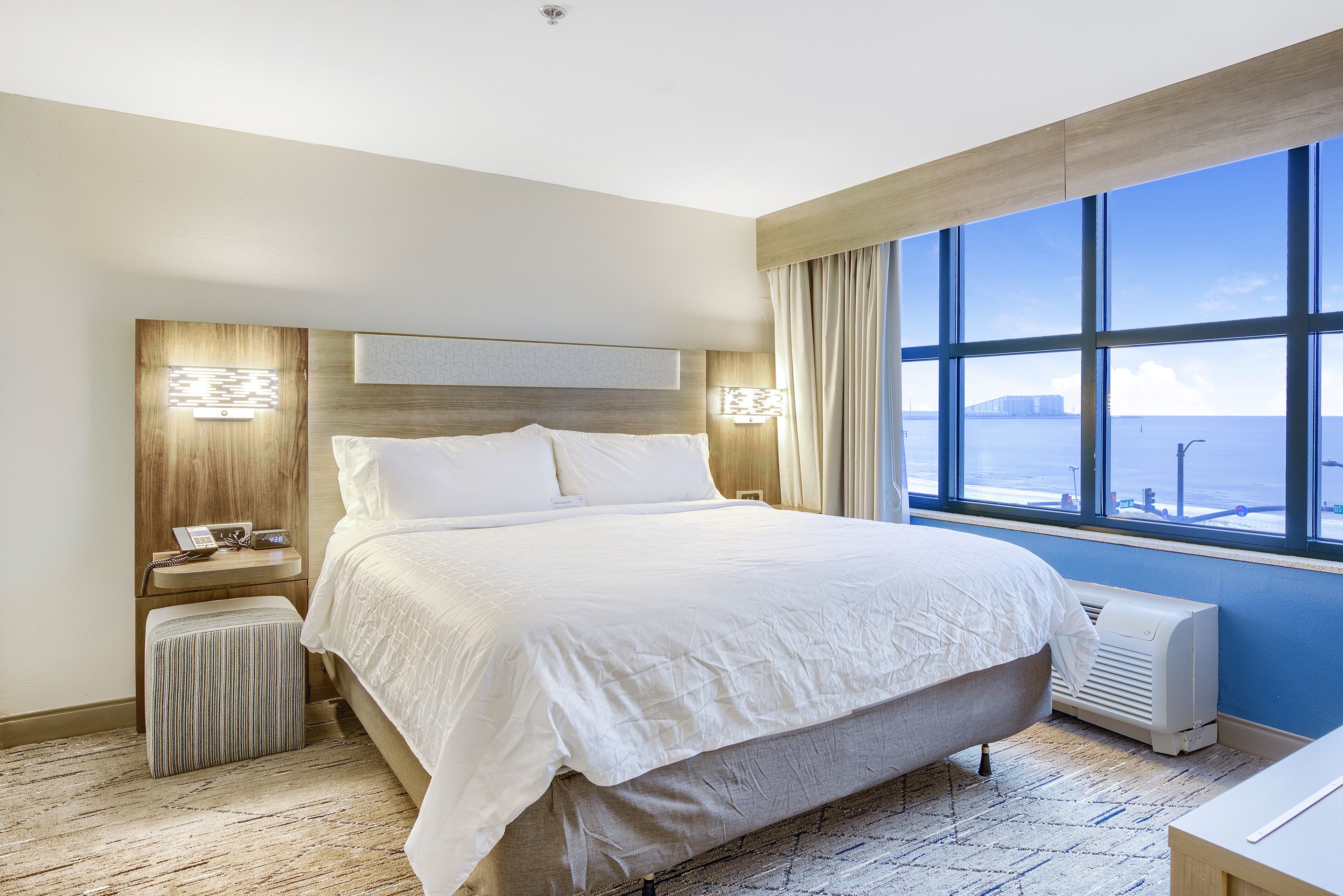Une chambre d’hôtel avec un grand lit, une table de chevet avec une lampe, un petit pouf et une vue sur la mer grâce à de grandes fenêtres.