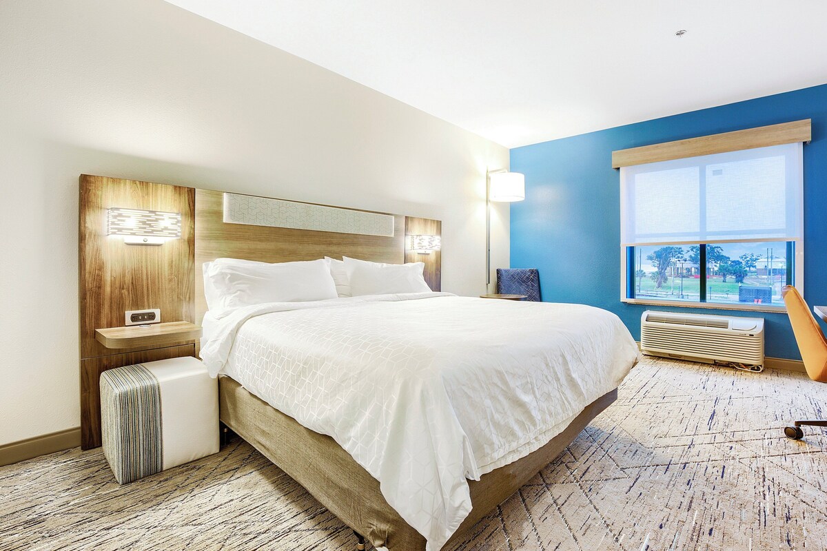 A modern hotel room with a large bed, a nightstand, a bedside table, a window with a view, and a wall-mounted heater.