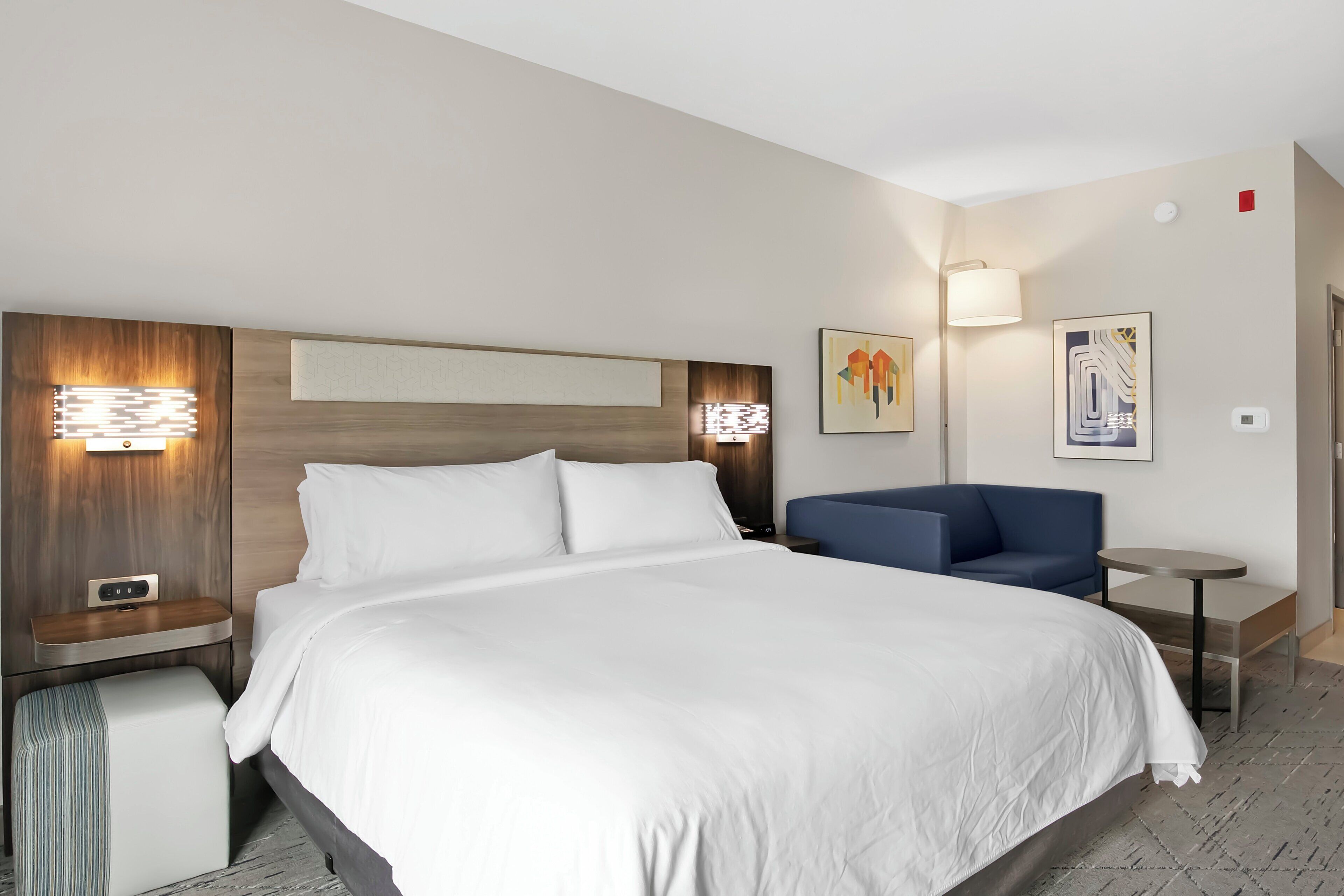 A hotel room with a large bed, a bedside table with a lamp, a small table, a blue sofa, and artwork on the walls.